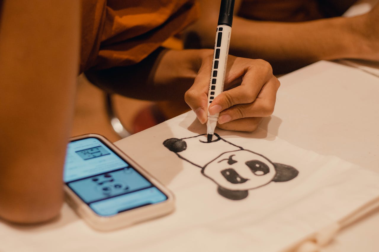 A person draws a panda on paper while referencing a smartphone for guidance.